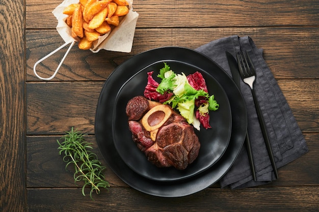 Bifes Osso Buco Carne fatiada grelhada Osso Buco ou bife de carne de porco com guarnecido com salada e batatas fritas na placa de mármore preto sobre fundo de madeira velho Vista superior Mock up