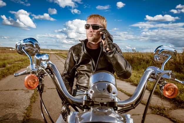 Biker hablando en un teléfono inteligente. Biker hombre vestido con una chaqueta de cuero y gafas de sol sentado en su motocicleta.