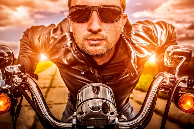 Biker hombre vestido con una chaqueta de cuero y gafas de sol sentado en su motocicleta mirando la puesta de sol. Filtro aplicado en postproducción.