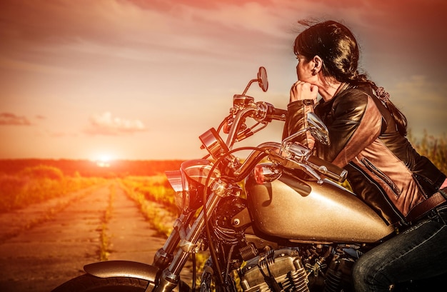 Biker-Mädchen in einer Lederjacke auf einem Motorrad, das den Sonnenuntergang betrachtet.