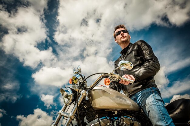 Foto biker-mann mit lederjacke und sonnenbrille sitzt auf seinem motorrad und blickt auf den sonnenuntergang.