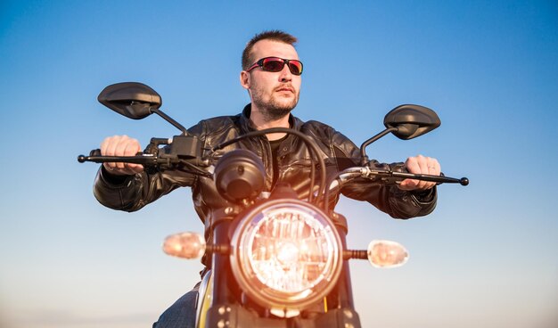 Biker-Mann mit Lederjacke und Sonnenbrille sitzt auf seinem Motorrad und blickt auf den Sonnenuntergang.