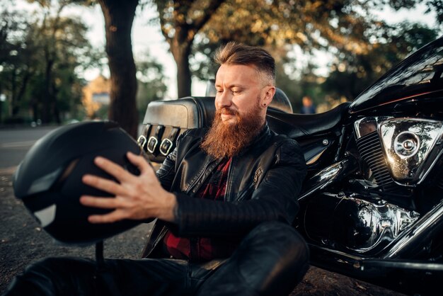 Biker mit Helm in den Händen sitzt auf dem Boden gegen seinen Hubschrauber.