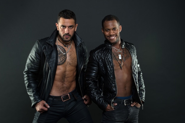 Biker mit sexy nackten Oberkörpern, Rebellenkonzept. Brutale Männer tragen Lederjacken auf schwarzem Hintergrund isoliert. Models in trendigen männlichen Accessoires. Afrikanische und hispanische Männer mit tätowierten Brüsten.