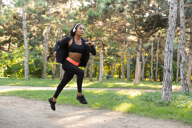 Bild der athletischen Frau 20s, die schwarzen Trainingsanzug und Kopfhörer tragen, während sie durch grünen Park laufen