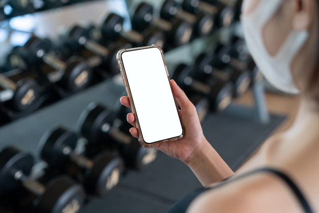 Bild der Frau, die Gesichtsmaske und ihre Hand hält, die Handy mit weißem Bildschirm am Fitnessstudio hält.