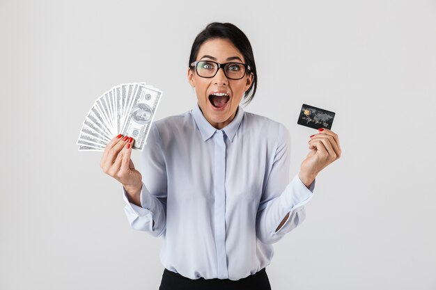 Bild der glücklichen Bürofrau, die Brillen trägt, die Bündel des Geldes und der Kreditkarte halten, lokalisiert über weißer Wand