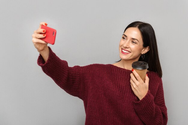 Bild der hübschen jungen Frau, die im burgunderfarbenen Pullover gekleidet ist, nehmen selfie durch Handy lokalisiertes Kaffeetrinken.