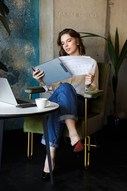 Bild der hübschen schönen jungen Frau, die im Café drinnen arbeitet, arbeitet mit Laptop, der Dokumente hält.