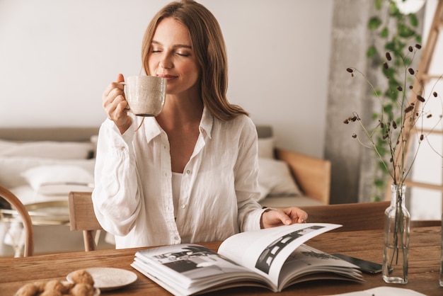 Bild der jungen erstaunlichen hübschen Frau sitzt drinnen im Büro und liest Zeitschrift, die Kaffee trinkt.