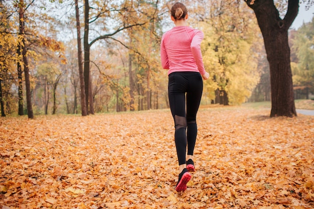 Bild der jungen Frau, die im Park läuft. Draußen ist es kalt. Orange Blätter liegen auf dem Boden. Junge Frau beim Joggen. Sie rennt alleine.