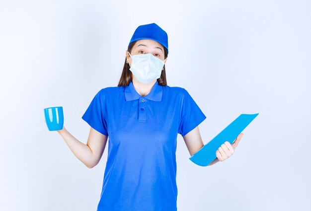 Bild der jungen Frau in medizinischer Maske mit blauem Ordner und Tasse.