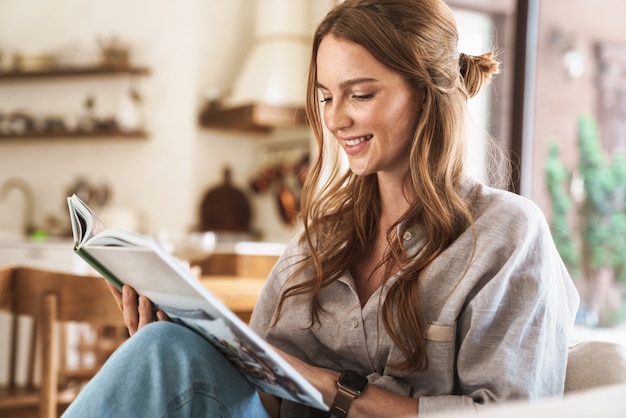 Bild der jungen hübschen schönen positiven optimistischen Rothaarigefrau, die zuhause zu Hause sitzt und ein Buch oder eine Zeitschrift liest.