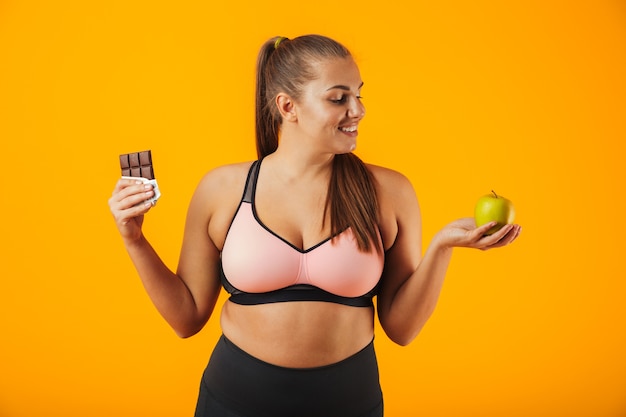 Bild der jungen übergewichtigen Frau im Trainingsanzug, der Schokoriegel und Apfel in beiden Händen hält, lokalisiert über gelbem Hintergrund