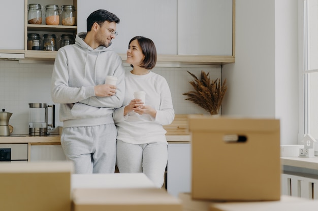 Bild der netten Frau und des Mannes haben angenehmes Gespräch während der Kaffeepause, tragen zufällige Kleidung, ziehen in neue Wohnung um, werfen mit moderner versorgter Küche auf, packen Pappschachteln mit Haushaltsmaterial aus