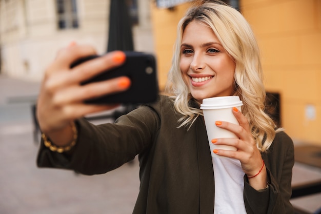 Bild der reizenden Frau, die Kaffee zum Mitnehmen in Pappbecher hält und Selfie nimmt, während durch Stadtstraße geht