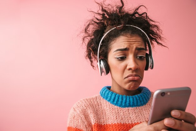 Bild der schockierten unzufriedenen schönen jungen afrikanischen Frau, die isolierte hörende Musik mit Kopfhörern unter Verwendung des Handys aufwirft.