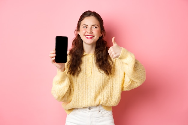 Bild der schönen Frau, die lacht und beiseite schaut, während sie eine App bewertet, die Daumen hoch und leeren Telefonbildschirm zeigt, der Online-Promo empfiehlt, der gegen rosa Wand steht