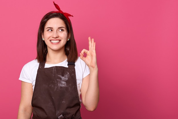 Bild der schönen glücklichen jungen Frau, die Küchenschürze und weißes T-Shirt trägt