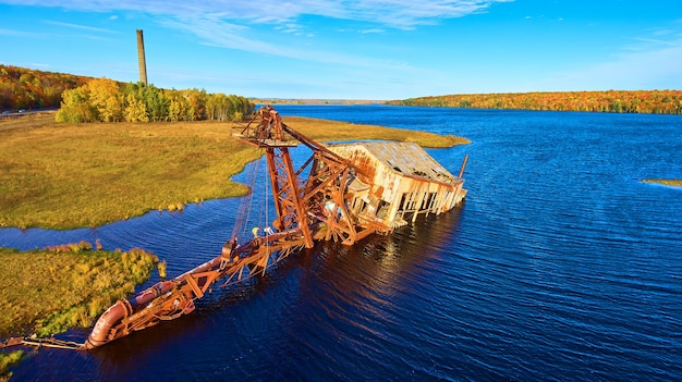 Bild des alten versunkenen Baggers in einem See zur goldenen Stunde mit blauem Himmel und einem Silo im Hintergrund mit Herbstbäumen