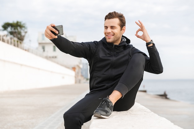 Bild des athletischen Mannes 30s in der schwarzen Sportbekleidung, Selfie-Foto auf Handy machend, während auf Promenade am Meer sitzend