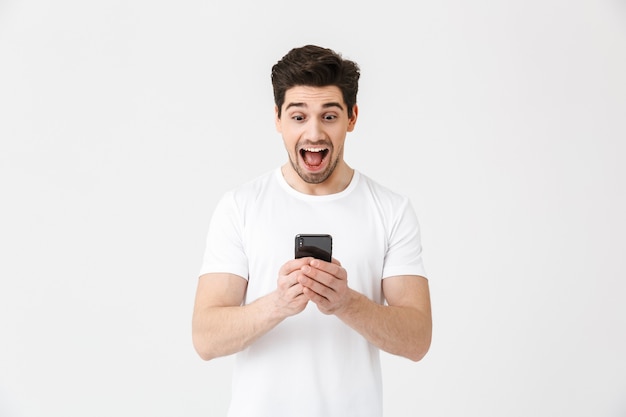 Bild des aufgeregten glücklichen jungen Mannes, der lokalisiert über weißer Wand mit Handy aufwirft.