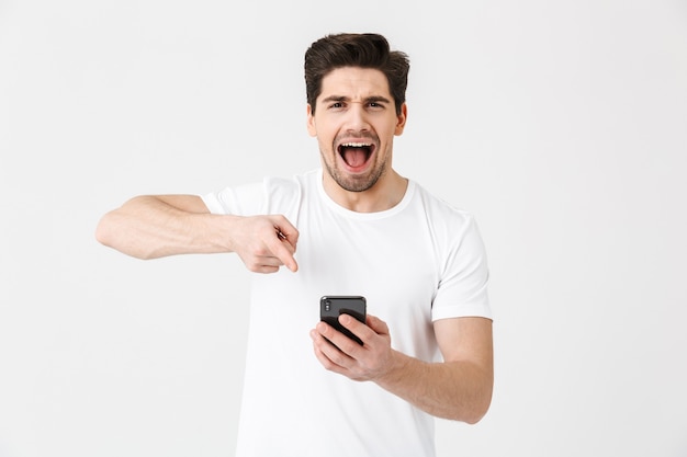 Bild des aufgeregten glücklichen jungen Mannes, der lokalisiert über weißer Wand mit Handy aufwirft.