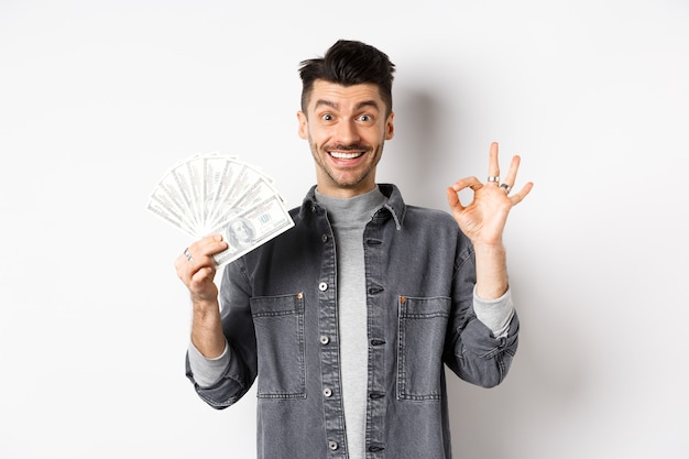 Bild des aufgeregten Mannes, der Dollarnoten hält und okay Zeichen mit glücklichem Gesicht zeigt, Geld verdient, auf weißem Hintergrund stehend.