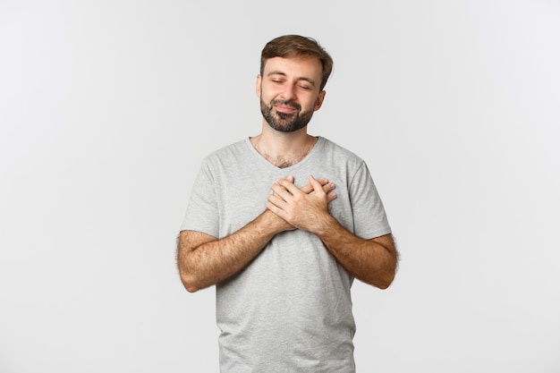 Bild des berührten bärtigen Mannes im einfachen T-Shirt, schließen Augen und Händchenhalten am Herzen