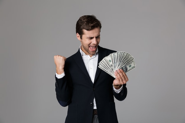 Bild des ekstatischen Geschäftsmannes 30s im Anzug lächelnd und hält Fan von Geldbanknoten, lokalisiert über graue Wand