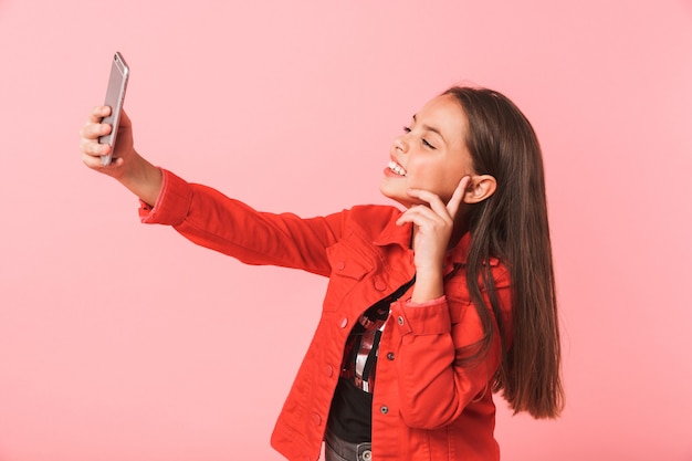 Bild des entzückenden jugendlich Mädchens in der lässigen Einnahme Selfie auf Handy während des Stehens, lokalisiert über rote Wand