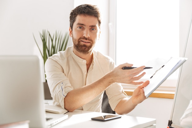 Bild des erfolgreichen Büro-Mannes der 30er Jahre, der weißes Hemd unter Verwendung von Laptop- und Papierdokumenten trägt, während am modernen Arbeitsplatz arbeitend