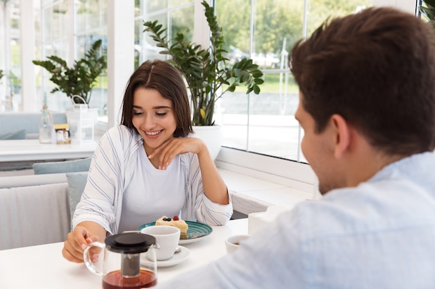 Bild des erstaunlichen jungen liebenden Paares, das im Café sitzt, essen Desserts und trinkt Tee, der miteinander spricht.