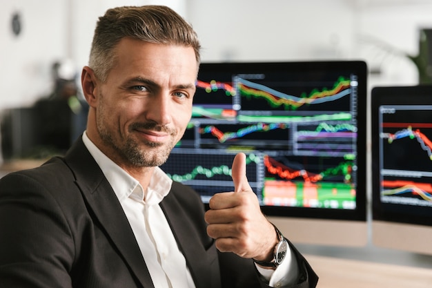 Bild des europäischen Geschäftsmannes 30s tragen Anzug, der im Büro am Computer mit Grafiken und Diagrammen am Bildschirm arbeitet