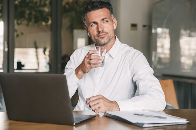 Bild des europäischen Geschäftsmannes der 30er Jahre, der weißes Hemd und Ohrhörer trägt, die am Tisch im Büro sitzen und Wasser aus Glas trinken, während am Laptop arbeiten