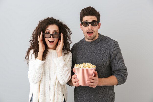 Bild des freudigen Mannes und der Frau, die 3D-Schutzbrillen tragen, die Eimer mit Popcorn halten, lokalisiert über graue Wand