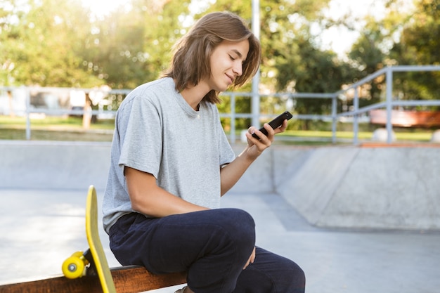Bild des fröhlichen jungen Skater-Kerls sitzen im Park mit Skateboard unter Verwendung des Mobiltelefons.