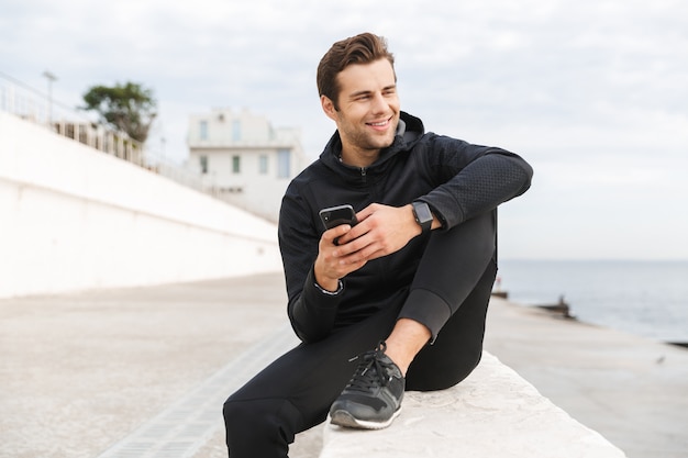 Bild des fröhlichen Sportlers 30s in der schwarzen Sportbekleidung, die Smartphone beim Sitzen auf Promenade am Meer verwendet