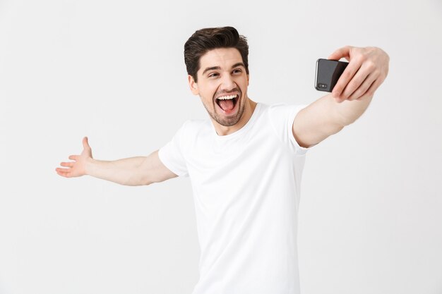 Bild des glücklichen emotionalen jungen Mannes, der lokalisiert über weißer Wand aufwirft, nehmen selfie durch Handy.