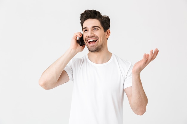 Bild des glücklichen jungen Mannes, der lokalisiert über der weißen Wand aufwirft, die durch Handy spricht.