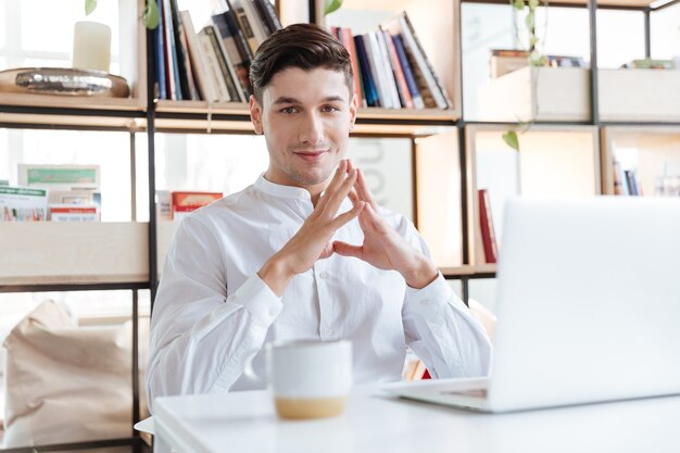 Bild des glücklichen Mannes gekleidet im weißen Hemd, das Kaffee trinkt und Laptop-Computer verwendet. Coworking. Blick in die Kamera.