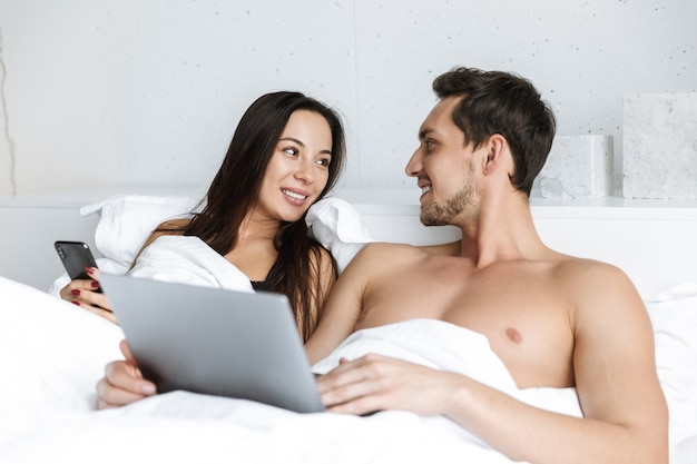 Bild des glücklichen Paares unter Verwendung des Laptops und des Smartphones, während im Bett zu Hause oder in der Hotelwohnung liegend