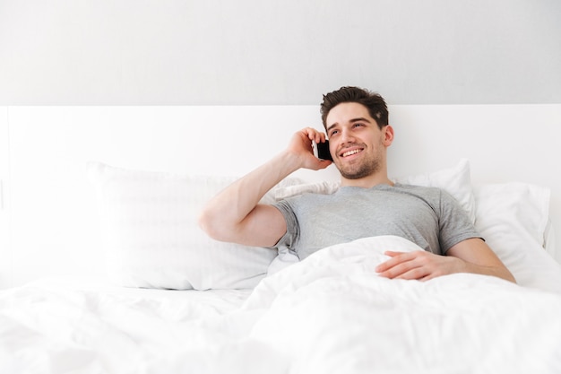 Bild des glücklichen wachen Mannes im T-Shirt lächelnd, während sie allein im Bett mit weißer Wäsche liegen und angenehmen mobilen Anruf haben