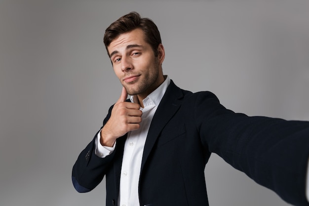Bild des gutaussehenden Mannes 30s in der formellen Anzugaufstellung beim Aufnehmen des Selfie-Fotos, lokalisiert über graue Wand