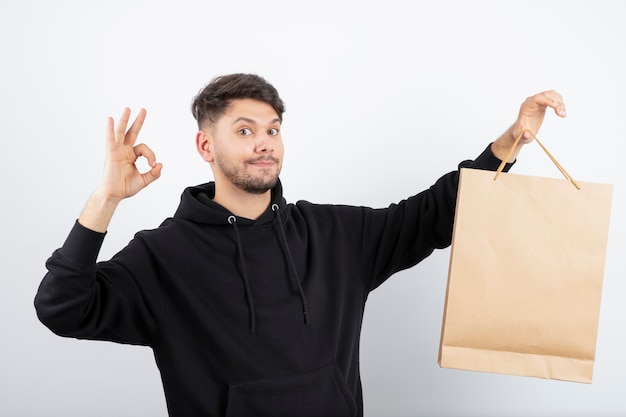 Bild des gutaussehenden mannes im schwarzen kapuzenpulli, der basteltasche hält, die ok zeichen gibt