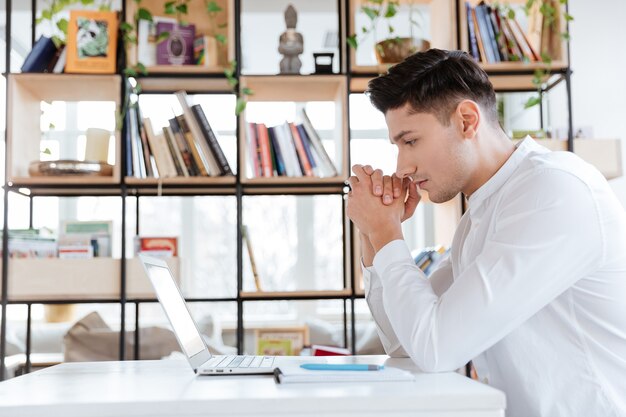 Bild des jungen ernsten Mannes im weißen Hemd mit Laptop-Computer gekleidet. Coworking. Computer betrachten.