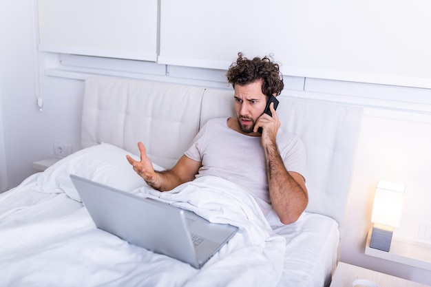 Bild des jungen kaukasischen Mannes hat Morgenkaffee, sitzt auf dem Bett, arbeitet an seinem Laptop und spricht mit seinem Handy, bereitet sich auf die Untersuchung vor, benutzt Laptop-Computer zum Surfen im Internet.