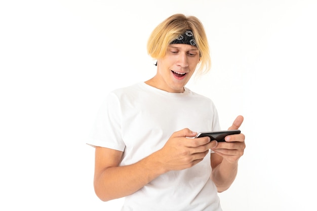 Bild des jungen kaukasischen Mannes mit blonden Haaren in kurzen Hosen und weißem T-Shirt, das Internet mit seinem Telefon serfing