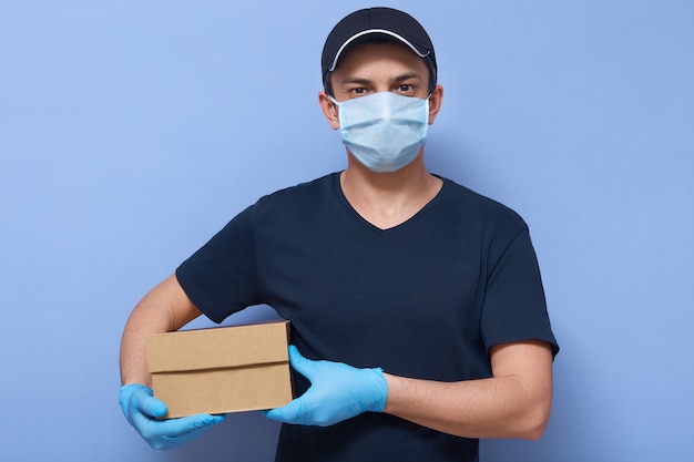 Bild des jungen Kuriers mit der Kartonschachtel in den Händen, die direkt auf die Kamera schauen, die über der blauen Wand lokalisiert wird, liefert Online-Shop-Mitarbeiter Bestellung an Kunden, besonderen Service während der Quarantäne.