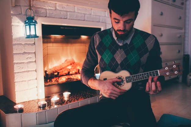 Bild des jungen Mannes, der am Kamin sitzt und Ukulele spielt